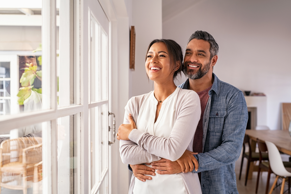 Smiling couple embracing beside a sunny window at home