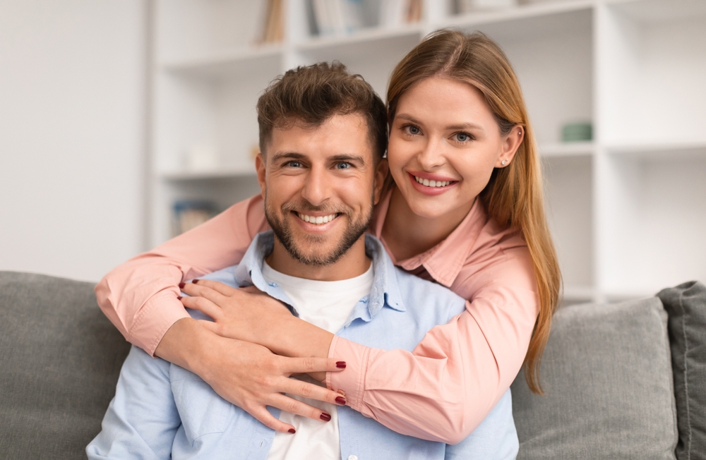 Smiling woman hugging a smiling man from behind on a sofa